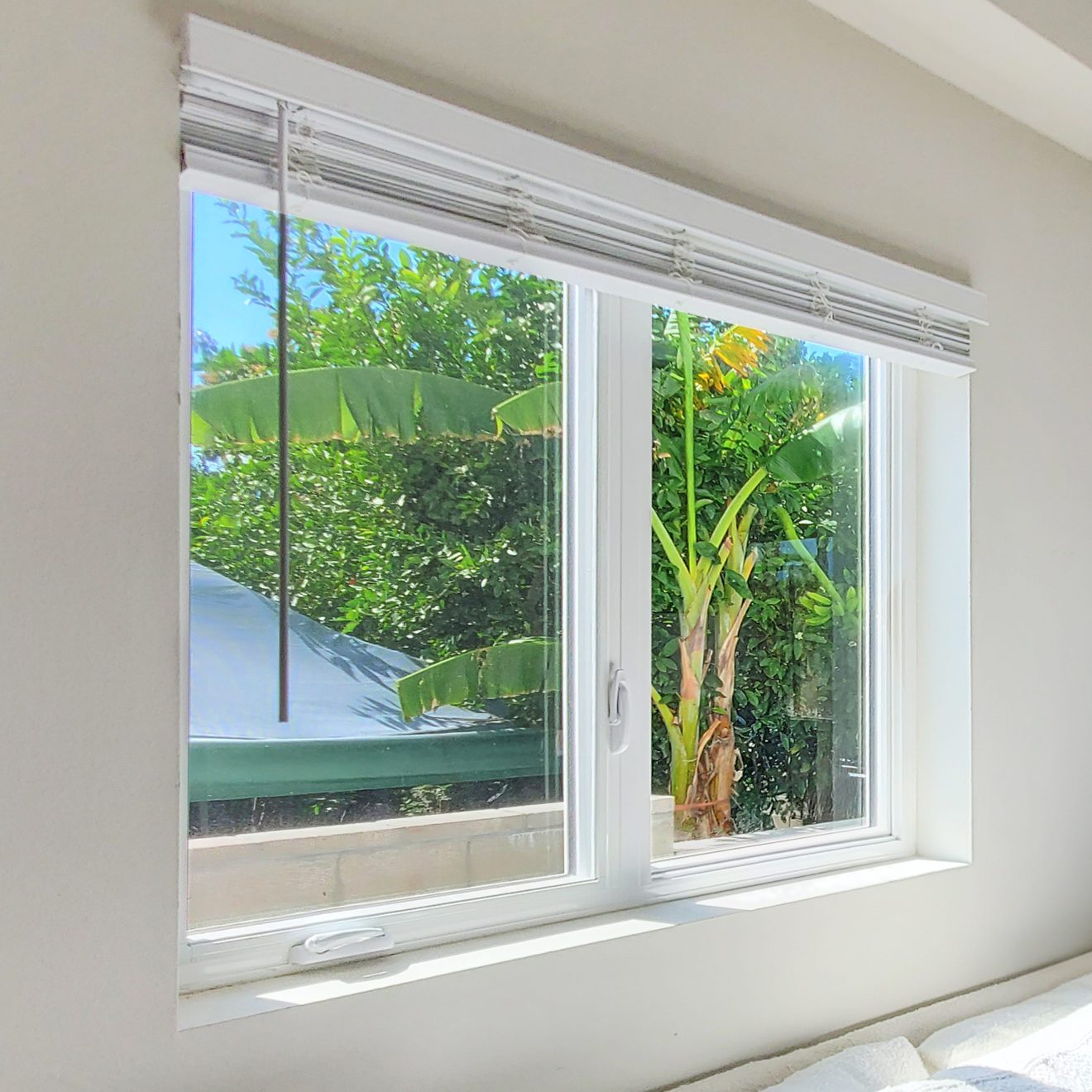 A white GS Series double-hung window with blinds partially open, revealing a view of lush greenery and a blue sky.